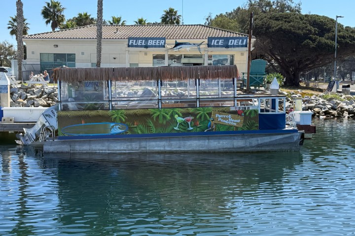 Tiki-themed boat docked near building with 'Fuel Beer Ice Deli' signs and palm trees.